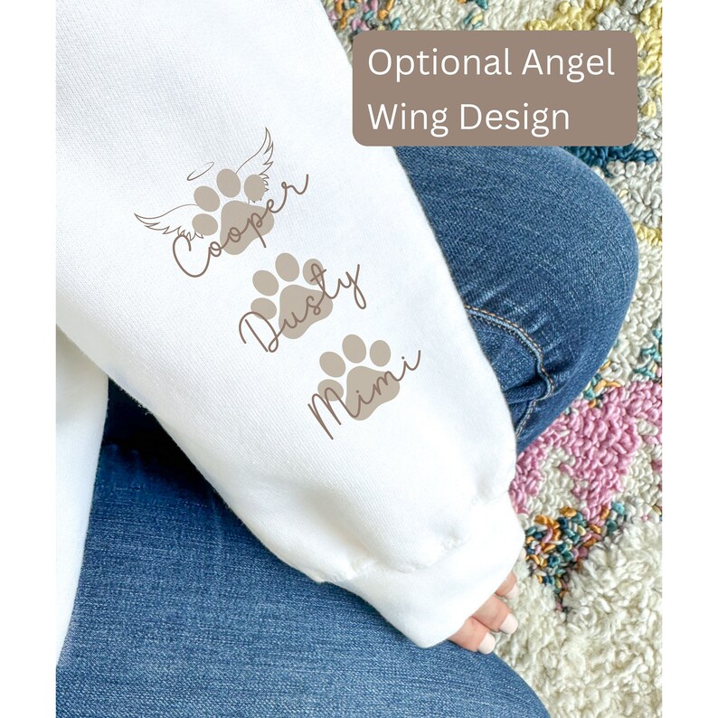 May include: White sweatshirt with brown paw prints and names "Cooper", "Dusty", and "Mimi" printed on the sleeve. The text "Optional Angel Wing Design" is printed on the top of the sleeve.