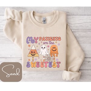Może przedstawiać: Beżowa bluza z napisem "My Patients are the Sweetest" w stylu retro, z grafiką cukierków kukurydzianych, ducha i lizaka z dyni. Projekt ma motyw Halloween.