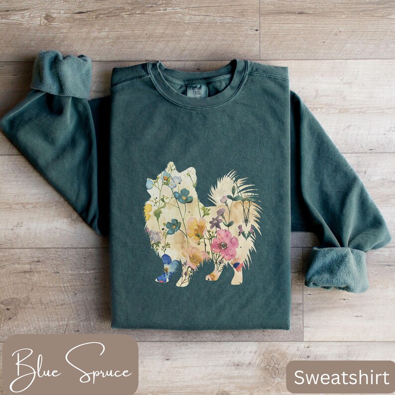 May include: A teal-colored sweatshirt featuring a floral Pomeranian design. The dog silhouette is filled with colorful flowers. The words "Blue Spruce" are at the bottom left, and "Sweatshirt" is at the bottom right.