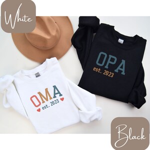 May include: Two sweatshirts, one white and one black, with text. The white sweatshirt says "OMA est. 2023" in colorful letters with two red hearts. The black sweatshirt says "OPA est. 2023" in teal and orange letters. A tan hat is visible.