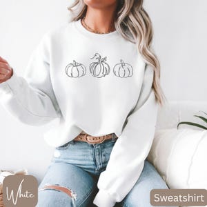May include: White sweatshirt with a line drawing of three pumpkins. The pumpkins are in a row, with the middle one having a stem and leaves. The word "Sweatshirt" is in a brown rectangle in the bottom right corner.
