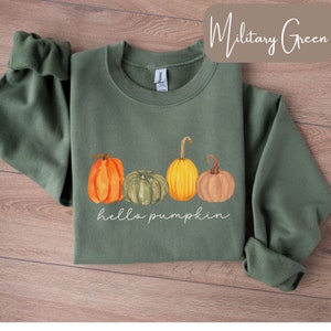 May include: Military green sweatshirt with a watercolor design of four pumpkins in orange, green, and yellow. The words "hello pumpkin" are written in white script below the pumpkins. The sweatshirt is on a wooden surface.