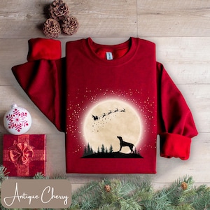 May include: A burgundy sweatshirt with a graphic of a dog silhouetted against a full moon with Santa's sleigh. The design includes a forest silhouette and falling stars. The text "Antique Cherry" is at the bottom.