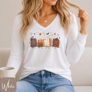 Op de afbeelding: Een wit shirt met lange mouwen en V-hals voorzien van een grafische print van antropomorfe boeken, een pompoenmok en herfstthema-iconen zoals vleermuizen, spinnenwebben en bladeren. De boeken zijn gelabeld met Moonlight Stories en Enchanted Library.