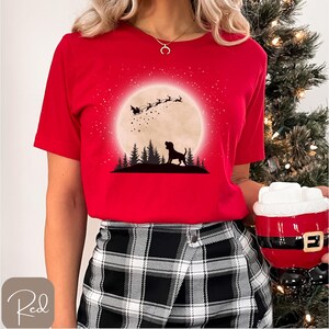May include: A red t-shirt featuring a silhouette of a dog in front of a full moon with Santa's sleigh and reindeer. The design includes a forest silhouette and a starry sky. The word "Red" is in a cursive font in the bottom left corner.