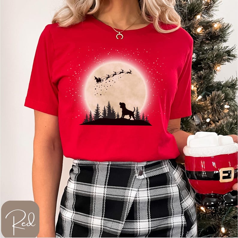 May include: A red t-shirt featuring a silhouette of a dog in front of a full moon with Santa's sleigh and reindeer. The design includes a forest silhouette and a starry sky. The word "Red" is in a cursive font in the bottom left corner.