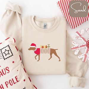 Könnte beinhalten: Cremefarbenes Sweatshirt mit einem braunen Hund, der einen Weihnachtsmannhut und einen roten Pullover trägt. Der Hund hat das Wort "HOHOHO" auf der Seite aufgedruckt, mit bunten Geschenkboxen darüber. Ein festliches Weihnachtsdesign.