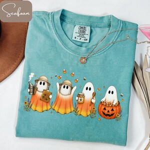 May include: A seafoam green t-shirt featuring a graphic of four ghosts dressed as beekeepers. The ghosts are surrounded by candy corn, bees, and flowers. One ghost holds a smoker and honeycomb, another a honey jar and dipper, and one sits in a pumpkin.