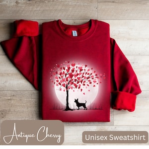 Puede incluir: Sudadera unisex roja con un gráfico de una silueta de un perro de pie debajo de un árbol con hojas en forma de corazón. El árbol está frente a una luna llena. El texto "Antique Cherry" está en la parte inferior izquierda de la imagen y "Unisex Sweatshirt" en la parte inferior derecha.