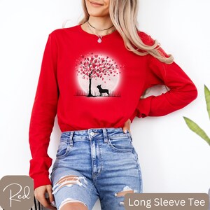 May include: Red long-sleeve t-shirt with a graphic of a silhouette of a dog standing under a tree with pink hearts for leaves. The tree is in front of a full moon.