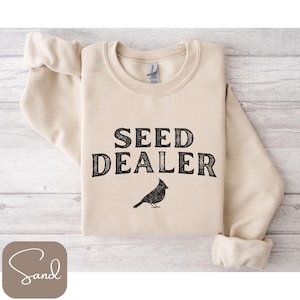 Op de afbeelding: Een zandkleurige sweatshirt met de tekst "SEED DEALER" in zwart, boven een zwarte vogelillustratie. De sweatshirt is netjes op een licht houten oppervlak gevouwen.