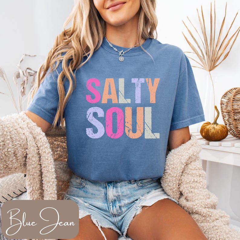 May include: A blue jean colored t-shirt with the words "SALTY SOUL" in colorful, distressed lettering. The shirt is short-sleeved and has a relaxed fit. The image also shows a beige sweater and denim shorts.