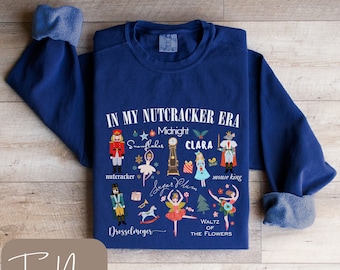 Puede incluir: Una sudadera unisex azul marino con el texto "IN MY NUTCRACKER ERA" e ilustraciones temáticas de El Cascanueces. El diseño incluye personajes como el Cascanueces, Clara y el Rey Ratón. La sudadera es "True Navy".