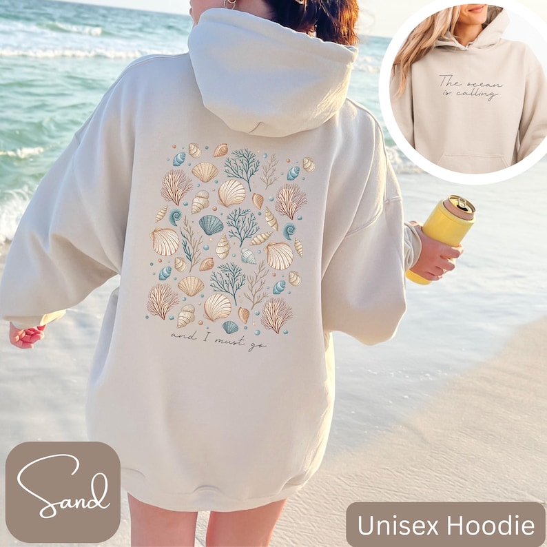 K&ouml;nnte beinhalten: Ein beiger Unisex-Hoodie mit einem Muschel- und Korallendesign auf der R&uuml;ckseite. Der Text "and I must go" ist unter dem Design gedruckt. Die Vorderseite des Hoodies hat den Text "The ocean is calling" auf der Tasche.