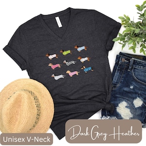 May include: Dark grey heather unisex V-neck t-shirt with a pattern of dachshunds in sweaters. The dachshunds have different colored sweaters. The shirt is displayed with a straw hat and denim shorts.