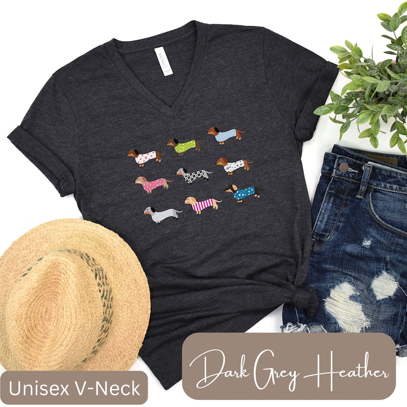 May include: Dark grey heather unisex V-neck t-shirt with a pattern of dachshunds in sweaters. The dachshunds have different colored sweaters. The shirt is displayed with a straw hat and denim shorts.