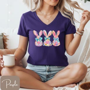 Op de afbeelding: Een paars V-hals T-shirt met drie aquarel konijn ontwerpen. Elk konijn heeft een strik, bloemmotieven en een hart. Het woord "Purple" staat in cursiefschrift linksonder.
