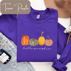 May include: A purple sweatshirt featuring a watercolor design of four pumpkins in shades of orange, green, and yellow. The words "hello pumpkin" are written in a script font below the pumpkins. The text "Team Purple" is in the upper left corner.