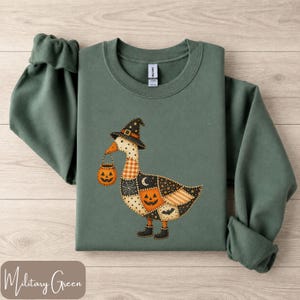May include: A military green crewneck sweatshirt featuring a graphic of a goose wearing a witch hat and boots. The goose has a patchwork design with orange, black, and white patterns, including pumpkins, stars, and a spiderweb, while holding a small orange jack-o'-lantern bucket.