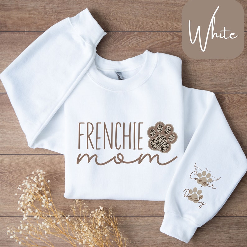 May include: White sweatshirt with the text "Frenchie mom" in brown lettering and a brown paw print with a leopard print design. The right sleeve has the text "Cooper & Dusty" in brown lettering with a paw print.
