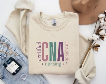 May include: A beige sweatshirt with the text "certified CNA nursing assistant" in colorful lettering. The sweatshirt is laid out on a white surface with a pair of jeans, a candle, and a hat.