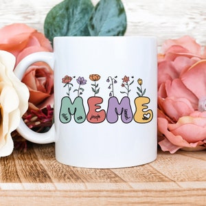 Może przedstawiać: Biały ceramiczny kubek z napisem "MEME" w kolorowych literach bąbelkowych, z których każda jest wazonem z innym kwiatem. Na kubku wydrukowano imiona "Olivia", "Eva", "Joseph" i "Samuel".