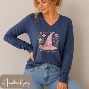 May include: A heather navy long-sleeve V-neck shirt featuring a central graphic of a pink floral patterned witch hat surrounded by small flowers and stars. The shirt is paired with blue denim jeans. The text Heather Navy is visible at the bottom.