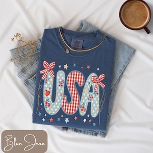 May include: A blue Comfort Colors t-shirt with the word "USA" in a patriotic design. The letters are decorated with floral patterns, gingham, and bows. The shirt is paired with blue jeans and a cup of coffee.