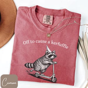 May include: A crimson short-sleeved t-shirt featuring a graphic of a raccoon wearing a party hat and riding a scooter. The text "Off to cause a kerfuffle" is printed in white above the raccoon. The shirt has a Comfort Colors brand label inside.