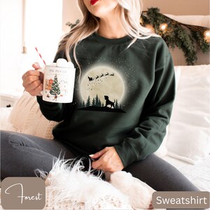 May include: Forest green sweatshirt featuring a silhouette of a dog, trees, and Santa's sleigh against a full moon. The sweatshirt has the word "Sweatshirt" printed on the bottom right corner.