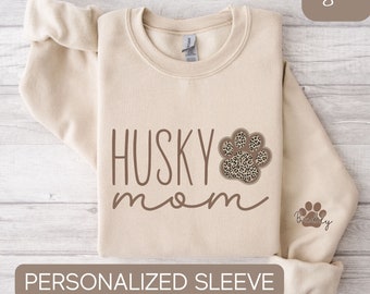 Op de afbeelding: Een beige sweatshirt met de tekst "HUSKY mom" in bruine letters. Naast de tekst staat een bruine pootafdruk met een luipaardprint. De mouw heeft een kleinere bruine pootafdruk met de tekst "Buddy" in bruine letters.