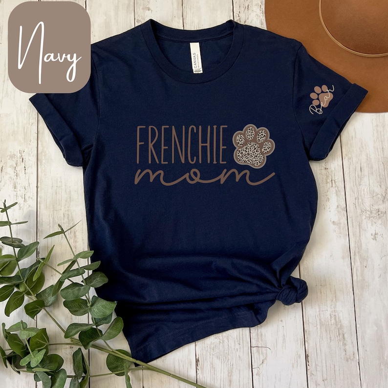 May include: Navy blue t-shirt with the text "Frenchie mom" in brown lettering and a brown paw print with a leopard print design. The paw print is on the left side of the shirt. The shirt has a small paw print on the right sleeve.
