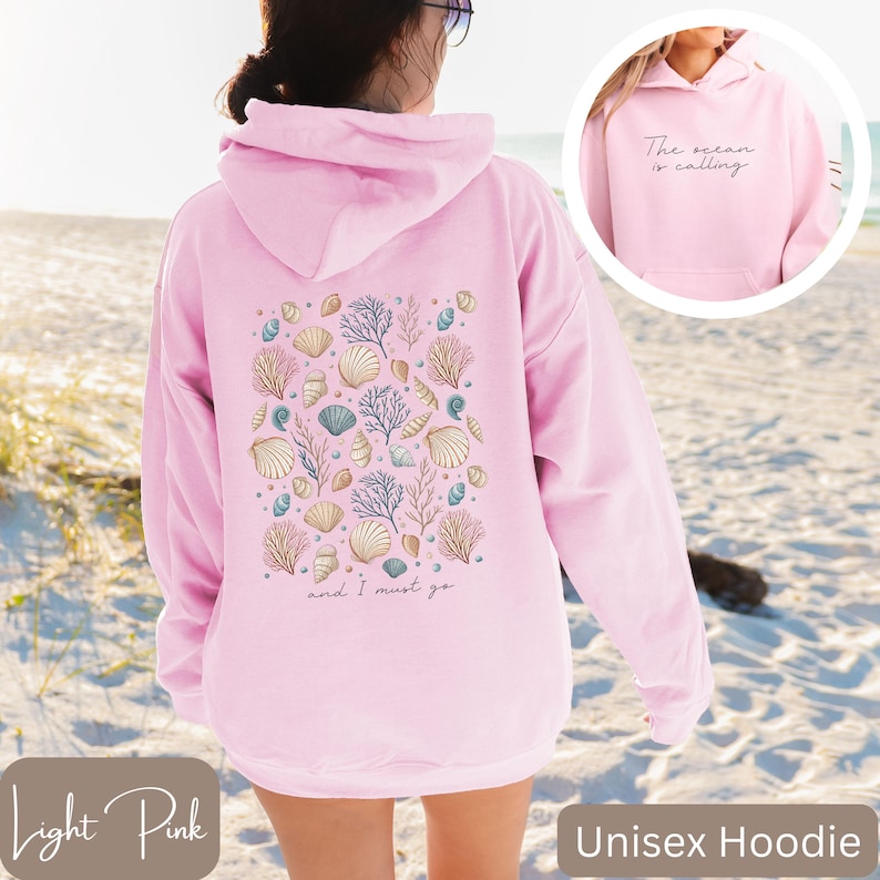 K&ouml;nnte beinhalten: Hellrosa unisex Hoodie mit einem Muschel- und Korallendesign auf der R&uuml;ckseite. Der Text "The ocean is calling and I must go" ist auf der R&uuml;ckseite des Hoodies aufgedruckt.