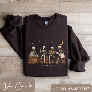 May include: A dark brown crewneck sweatshirt featuring a graphic print of four skeletons engaged in daily activities. The design includes a skeleton at a desk with a laptop, one standing, one holding a clipboard, and one walking with a tote bag.