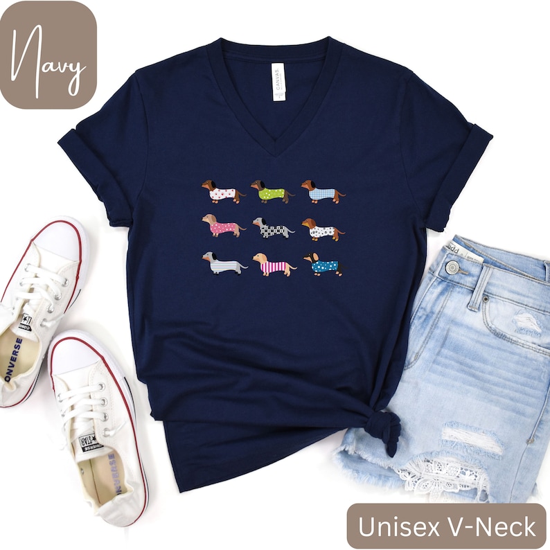 May include: Navy blue unisex V-neck t-shirt with a design of nine dachshunds in patterned coats. The dogs have different coat patterns and colours. The shirt is styled with denim shorts and white trainers.