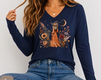 May include: A navy blue long-sleeve shirt with a V-neckline featuring a central graphic print of a figure wearing a wide-brimmed hat, surrounded by colourful wildflowers, foliage, a crescent moon, and small stars against a dark blue background.