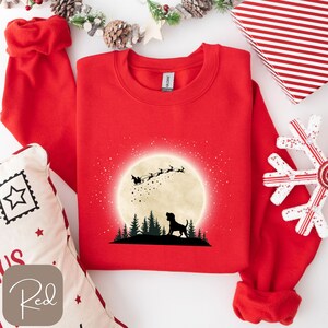 May include: A red sweatshirt featuring a graphic of a dog silhouetted against a full moon, with Santa's sleigh and reindeer flying above a forest. The word "Red" is printed on a small, square label.