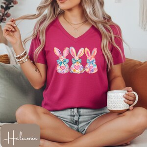 Op de afbeelding: Een magenta V-hals T-shirt met drie aquarel Paashaas ontwerpen. Elke konijn heeft een bloemenpatroon, een blauwe strik en een hart. Het shirt heeft korte mouwen en het woord "Heliconia" staat linksonder.