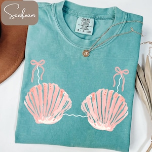 以下が含まれることがあります： シーフォームグリーンのTシャツ、貝殻のデザイン。ピンクの貝殻のイラストが2つ、白い線でつながっており、上にピンクのリボンが付いています。シャツにはComfort Colorsのタグと金のネックレスが付いています。