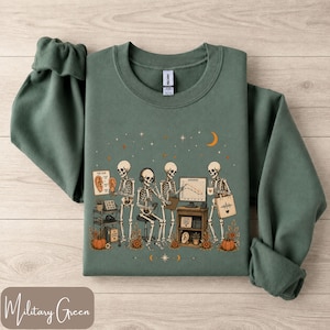 May include: A military green crewneck sweatshirt featuring a graphic print of four skeletons in a clinical setting. The illustration includes skeletons with headphones, a computer monitor displaying a graph, an ear diagram, and pumpkins.