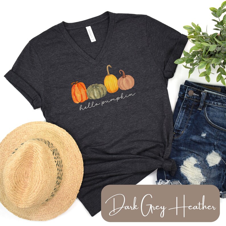 May include: Dark grey heather V-neck t-shirt featuring a watercolor design of four pumpkins in orange, green, and yellow. The words "hello pumpkin" are written in a script font below the pumpkins. A straw hat and denim shorts are also visible.