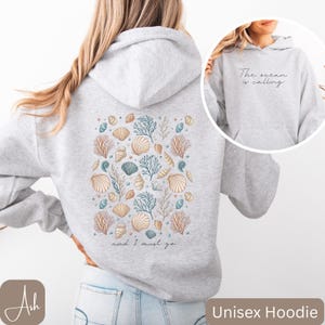 K&ouml;nnte beinhalten: Grauer Unisex-Hoodie mit einem Muschel- und Korallendesign auf der R&uuml;ckseite. Der Text "The ocean is calling and I must go" ist unter dem Design gedruckt.