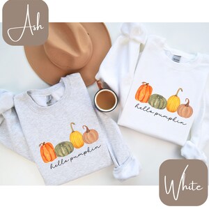 May include: Two sweatshirts, one ash gray and one white, each featuring a watercolor design of pumpkins in orange, green, and yellow. The phrase "hello pumpkin" is written in a script font below the pumpkins. A brown hat and a mug of coffee are also visible.