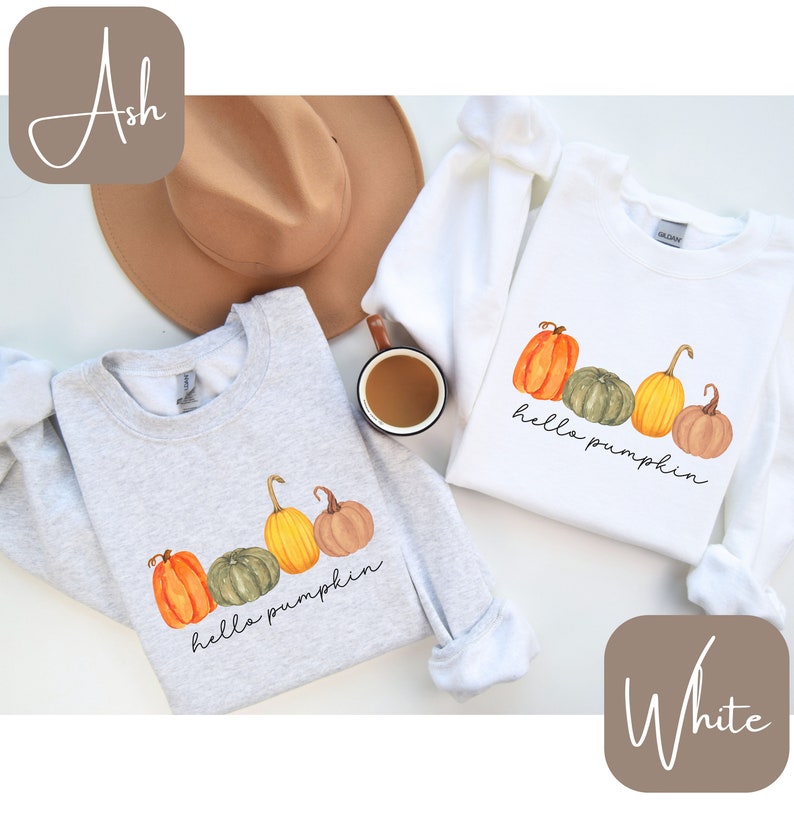 May include: Two sweatshirts, one ash gray and one white, each featuring a watercolor design of pumpkins in orange, green, and yellow. The phrase "hello pumpkin" is written in a script font below the pumpkins. A brown hat and a mug of coffee are also visible.