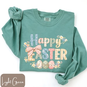 Puede incluir: Una camiseta de manga larga verde claro con las palabras "Happy Easter" en una fuente decorativa colorida. El diseño incluye un lazo, flores y huevos de Pascua. La camiseta está hecha de un material suave y es de ajuste cómodo.