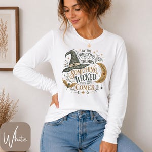 May include: A white long-sleeve shirt featuring a graphic print of a witch hat, a crescent moon, a bat, and the text "By the pricking of my thumbs, something wicked this way comes" in black, gold, and green tones with decorative botanical elements.