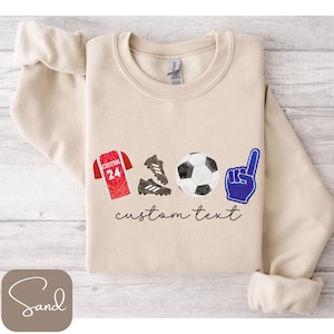 May include: Beige sweatshirt with soccer-themed graphics. Includes a red jersey with the number 24, soccer cleats, a soccer ball, and a blue foam finger. The words "custom text" are written in cursive.