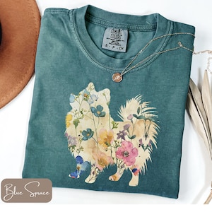 May include: Teal Comfort Colors t-shirt featuring a floral Pomeranian design. The dog silhouette is filled with colorful flowers in shades of pink, blue, yellow, and white. A gold necklace with a pendant is draped across the shirt.