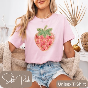 Puede incluir: Camiseta unisex rosa claro con un gran gráfico de fresa. La fresa tiene un tallo verde y está llena de un estampado floral en tonos rosas y rojos. El texto "Soft Pink" está en la parte inferior izquierda.