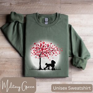 Op de afbeelding: Een unisex sweatshirt in militaire groene kleur met een silhouet van een poedel die onder een boom staat met roze harten als bladeren. De boom is omringd door een witte cirkel.